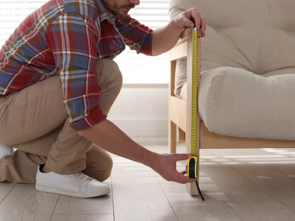 A man kneeling to measure the height of a wooden-framed armchair with a tape measure